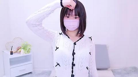 Media: Video of an Asian woman with short black hair, wearing a white lace cardigan with black buttons, a pink face mask, and a white surgical mask, adjusting her hair in a modern, minimalistic bedroom.