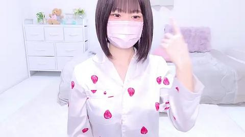 Media: Video of a young Asian woman with straight black hair, wearing a white shirt with pink strawberry patterns, a pink face mask, and a white surgical mask, in a brightly lit, minimalist bedroom with a white dresser and plush toys.