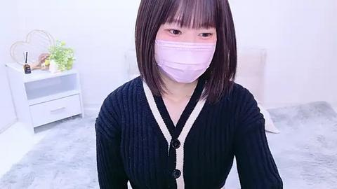 Media: Video of an East Asian woman with straight black hair and a light complexion, wearing a pink face mask, black cardigan, and white cardigan over it. She sits on a gray rug in a minimalist white room with a white dresser and potted plants.