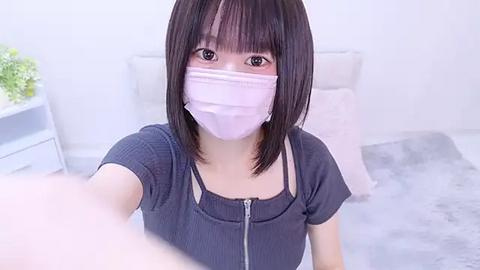 Media: A video of an East Asian woman with straight black hair and bangs, wearing a face mask, dark top, and light gray bedding.