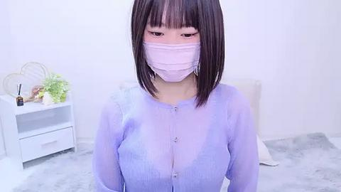 Media: Video of a young Asian woman with straight, shoulder-length black hair, wearing a see-through lavender button-up blouse, revealing her breasts. She has a face mask on and is in a minimalist white room with a white table and flowers.