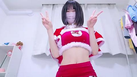 Media: Video of an East Asian woman with a short bob haircut, wearing a red Santa-themed crop top and pants, making peace signs, in a room with white curtains and clothes hanging on a rack.