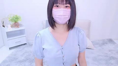 Media: Video of an Asian woman with straight black hair, light skin, wearing a light blue V-neck shirt, and a pink face mask in a minimalistic, white bedroom with a fluffy gray rug and white furniture.