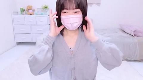 Media: Video of an East Asian woman with straight black hair, wearing a light pink face mask and light grey cardigan, standing in a minimalist, white-walled bedroom with a bed and white dresser.