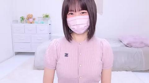 Media: Video of a young Asian woman with shoulder-length black hair, wearing a light pink face mask and matching ribbed top. Background includes a white dresser, plush toys, and a bed.