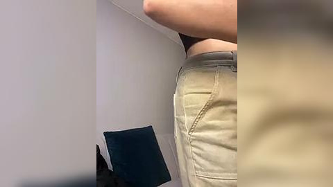 Media: Video of a person's upper torso and waist, wearing khaki shorts and a black shirt, standing in a dimly lit room with a dark green pillow and a white wall.