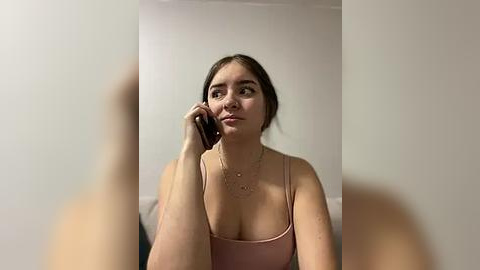 Media: Video of a fair-skinned woman with brown hair in a ponytail, wearing a pink spaghetti-strap tank top, talking on a smartphone, indoors against a plain white wall.