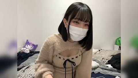 Media: Video of an East Asian woman with straight black hair, wearing a beige knit sweater with a lace-up front, a white face mask, and black headphones. She's in a cluttered bedroom with a purple stuffed cat and disorganized bed.