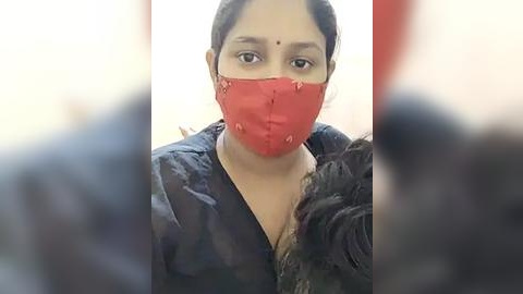 Media: Video of a young woman with medium brown skin, wearing a red mask, black blouse, and a floral jacket, taken indoors, blurred background.