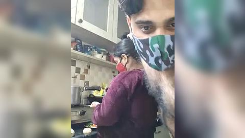 Media: Video of a man with a gray beard and a green face mask, cooking in a kitchen with beige tiles and cabinets.