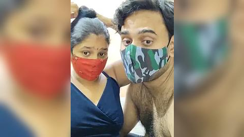 Media: Video of a South Asian couple in protective masks, with the woman in a red mask and blue top, and the man in a camo mask, taken indoors.