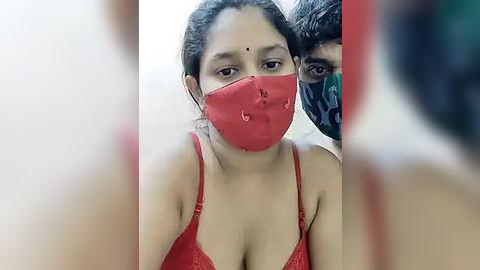 Media: Video of a South Asian woman with medium skin tone, wearing a red bra and red face mask, her dark hair pulled back, standing indoors with a man in the background partially visible.