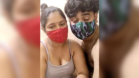 Media: Video of a South Asian woman with medium skin tone, wearing a red mask, gray tank top, and a man with dark skin and a camouflage mask, both sitting closely.