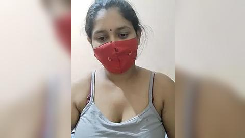 Media: Video of a South Asian woman with medium skin tone and dark hair tied back, wearing a red face mask and light gray spaghetti-strap tank top, revealing cleavage.