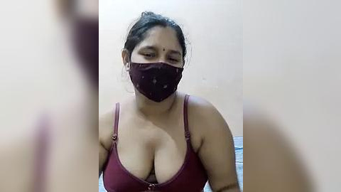 Media: A video of a South Asian woman with medium skin tone, wearing a maroon bra and face mask, her dark hair tied back, standing against a plain, light-colored wall.