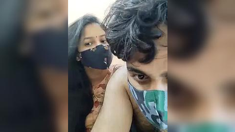 Media: Video of a man with curly dark hair and a woman with long black hair, both wearing black face masks, blurred in the foreground.