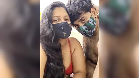 Media: Video of a couple in a close embrace, both wearing face masks and casual clothes. The woman has long black hair and a red top, while the man has curly black hair and a camouflage-patterned mask.