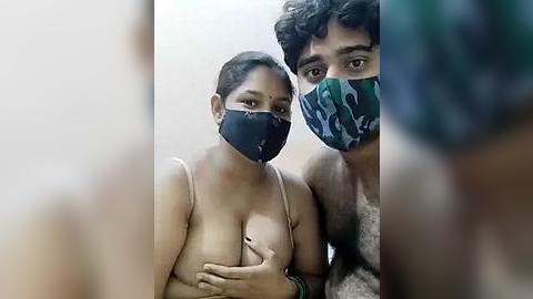 Media: Video of a young, light-skinned woman with medium-sized breasts wearing a blue face mask and beige bra, standing next to a man with curly hair in a green mask, both indoors.