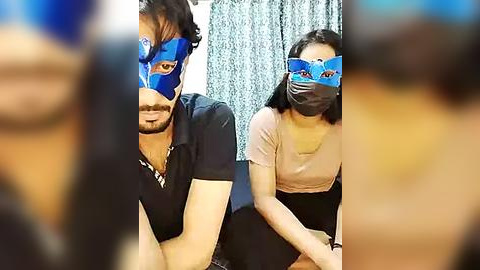 Media: Video of two Asian men in blue face masks, seated side-by-side in an office with light blue curtains. They wear black t-shirts and appear serious, with blurred figures in the foreground.