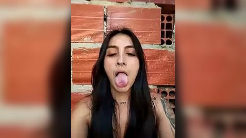 Media: Video of a young woman with long black hair, wearing a black top, sticking her tongue out against a red brick wall background.