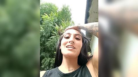 Media: Video of a smiling woman with long dark hair, wearing a black tank top, against a green foliage background. She has a nose ring and tattoos on her arm.