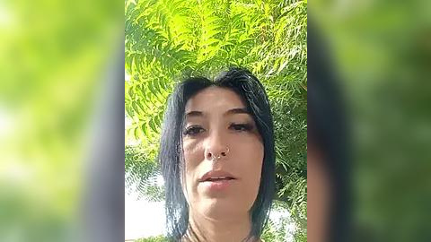 Media: Video of a young woman with short black hair, wearing a nose ring, and standing against a vibrant green background of ferns.