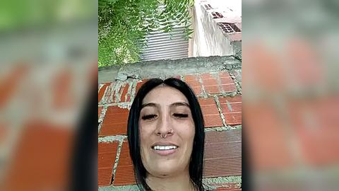 Media: Video of a young woman with straight black hair, a nose ring, and a broad smile, standing in front of a red brick wall with a green leafy tree in the background.