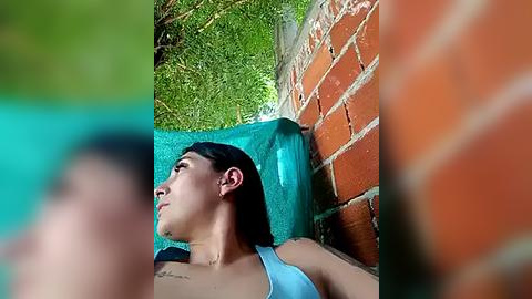 Media: Video of a young Asian woman with long black hair, wearing a blue tank top, sitting on a blue bench against a brick wall. She has a contemplative expression. The background features green foliage and a turquoise towel.