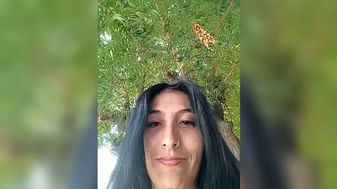 Media: Video of a young woman with long, dark hair, smiling, surrounded by lush green leaves. Her skin is light, and she has a septum piercing. A small, orange-brown lizard is visible on the foliage above her head.