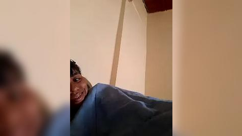 Media: Video of a joyful, dark-skinned woman peeking through a window, smiling widely. She wears a blue blanket, partially obscuring her face. The background shows a beige wall and part of a red ceiling.