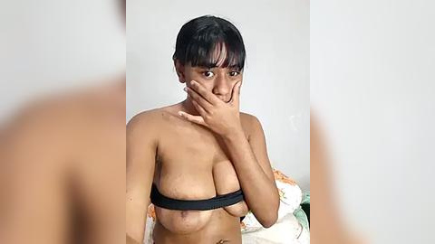 Media: Video of a topless Southeast Asian woman with medium brown skin, dark hair, and medium-sized breasts. She covers her mouth with her hand, standing against a plain white wall.