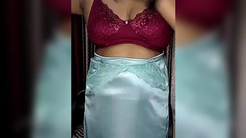 Media: Video of a woman in a maroon lace bra and light blue satin skirt, standing with arms raised, blurred background.