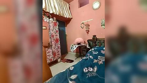 Media: A video of a small, cozy bedroom with pink walls, a floral-patterned curtain, a blue quilted bedspread, and a nightstand with a pink cushion.