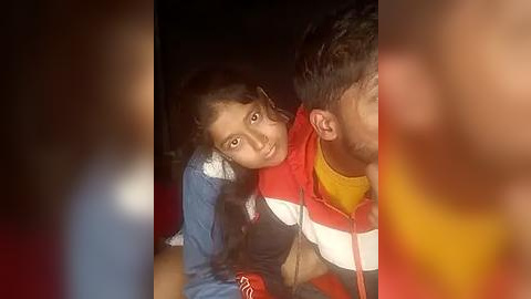 Media: Video of a young boy and girl sitting close together in a dimly lit room, with blurred faces and colorful clothing.