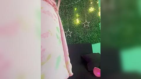 Media: Video of a person's lower body in a pink tie-dye dress, standing next to a green artificial turf wall with star lights and a black sofa.