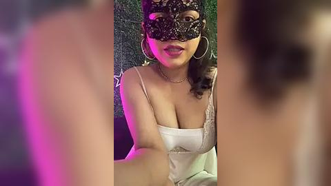 Media: A video shows a woman with light brown skin, wearing a black lace mask, white satin dress, and hoop earrings, sitting against a green wall.