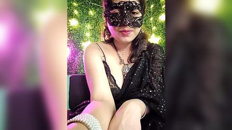 Media: Video of a woman with fair skin, dark hair, and a black lace mask, wearing a black sequined dress, sitting on a chair against a green leafy wall with yellow fairy lights.