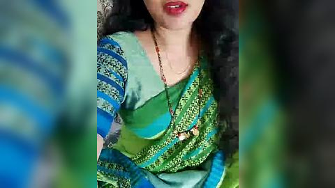 Media: A video of a woman with medium skin tone, wearing a vibrant, green and blue sari with intricate patterns, seated outdoors. Her black hair is wavy, and she has red lipstick.