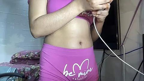 Media: Video of a light-skinned woman in a pink sports bra and matching shorts, holding a white cable, in a cluttered room with floral bedding and a TV.