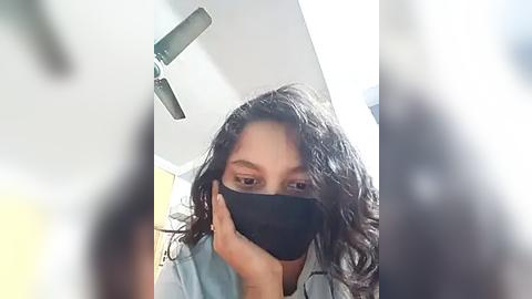 Media: A video of a young woman with medium skin tone, long dark hair, and a black face mask, taken indoors with bright natural light, wearing a light blue shirt.