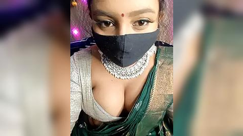 Media: Video of a South Asian woman with a fair complexion, wearing a traditional green and white sari, black face mask, and ornate silver necklace. Her dark eyes and red bindi are prominent.