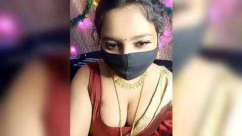 Media: Video of a South Asian woman with medium skin tone, wearing a red sari, black face mask, and gold necklace, with blurred background.