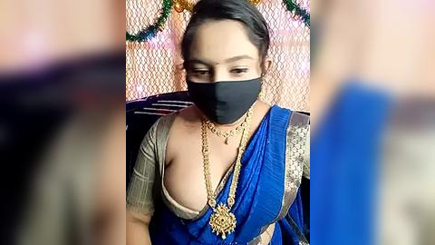 Media: A video of an Indian woman with light brown skin, wearing a black face mask, blue sari, gold jewelry, and a beige blouse, seated indoors with festive decorations in the blurred background.