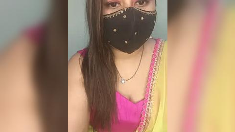 Media: Video of a young woman with long brown hair, wearing a black mask, pink and yellow saree, and a gold necklace, indoors against a blurred background.