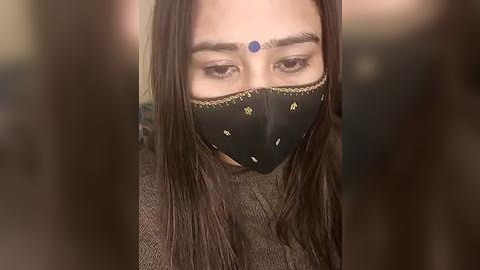 Media: Video of a woman with long, dark hair, wearing a black face mask with yellow floral patterns, and a blue bindi on her forehead. She has a neutral expression and is wearing a grey sweater.