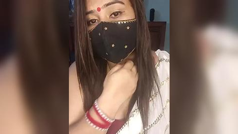 Media: Video of an Indian woman with fair skin, long dark hair, wearing a black face mask with red dot, traditional red bangles, and a white sari with gold embroidery, indoors with blurred background.