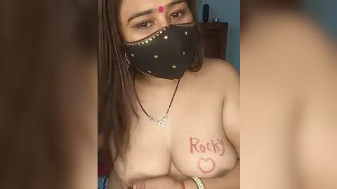 Media: Video of a topless woman with medium skin tone, brown hair, wearing a black mask, a red bindi, and \"Rocky\" written on her chest.