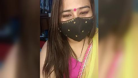 Media: Video of a woman with long brown hair, wearing a black mask with pink dots, a pink sari with yellow border, and a red dot on her forehead. Background blurred.