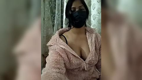 Media: Video of a woman with medium skin tone, wearing a pink fuzzy jacket, black face mask, and a black bra, taken indoors with patterned curtains in the background.