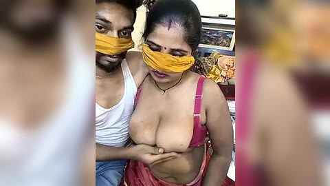 Media: Video of a South Asian couple in a domestic setting, man with medium build, dark skin, and short hair, wearing a white tank top, and woman with medium build, light brown skin, and medium-length black hair, wearing a pink bra and yellow mask, covering her face and chest.